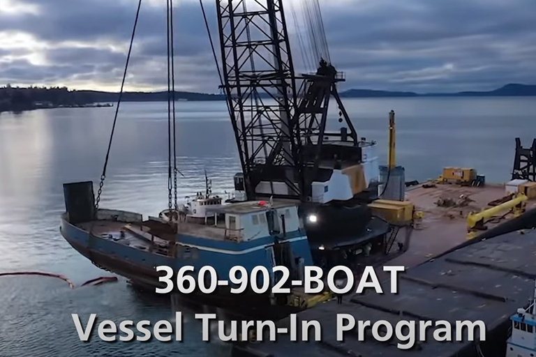 Screenshot from Washington State Department of Natural Resources derelict vessel video, showing a boat being lifted from the water with a crane, and a phone number, 360-902-BOAT, to call for the vessel turn-in program