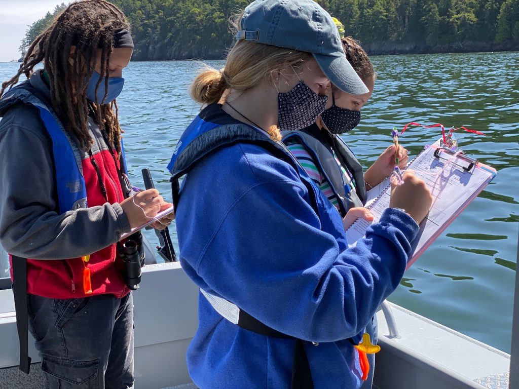 Salish Sea School students and faculty take notes and record data on the deck of a boat out on the water in Puget Sound