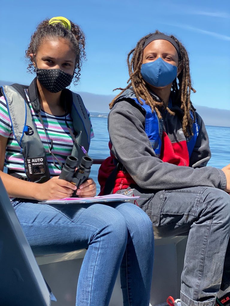 Two young students sit on a bench seat on a boat that's out on the water in the Puget Sound