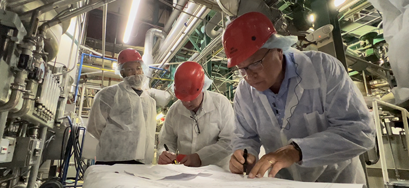 Three people in red hard harts look at detailed plans while standing inside a factory with machinery all around them.