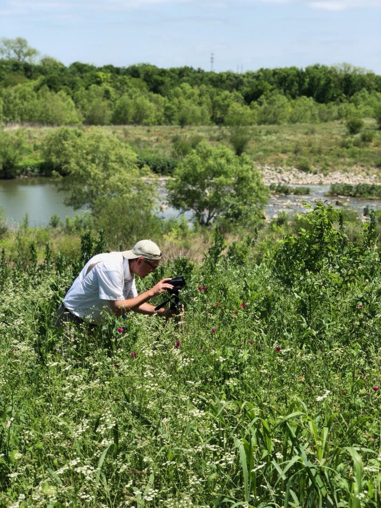 Photo of Steven Schauer, standing in vegetation next to the Mission Reach section of the San Antonio River, bent over a camera to capture video of the river.
