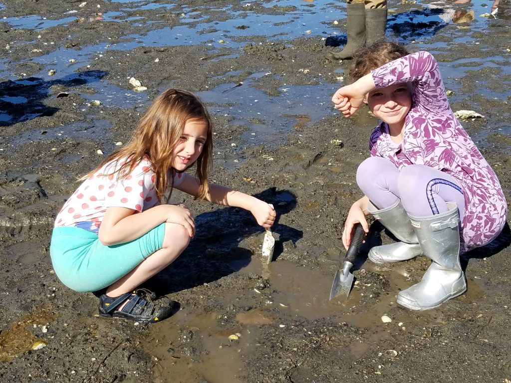 Two children with garden shovels pose for a picture while digging in the sand for shellfish.