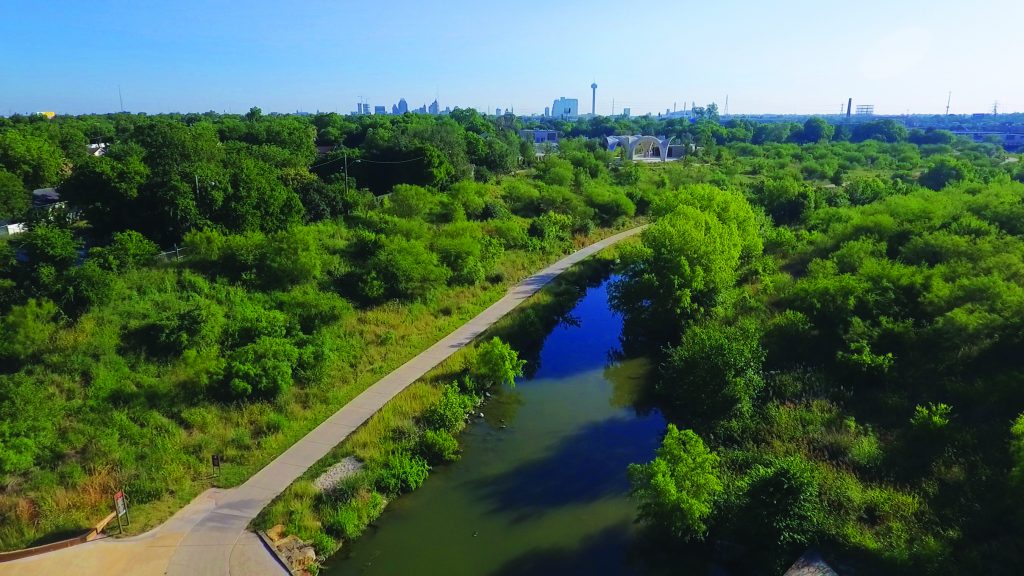 Aerial photo of the Mission Reach section of the San Antonio River, showing a walking path next to the river with the city of San Antonio in the distance.