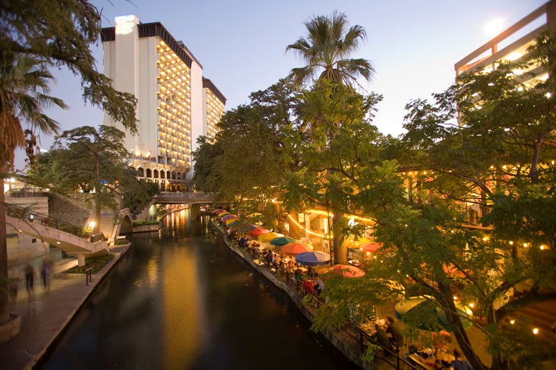 Photo of San Antonio River Walk, showing the San Antonio River flowing next to brightly lit buildings and cafes with patrons sitting under umbrellas.