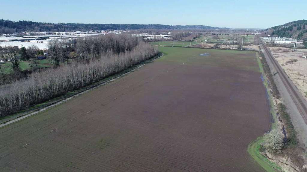 Aerial photo of the area near the White River that will be restored for side-channels and wetlands as habitat for salmon. The photo shows a large, flat grassy area next to a line of trees near the river, with large white buildings clustered together on the other side of the river.