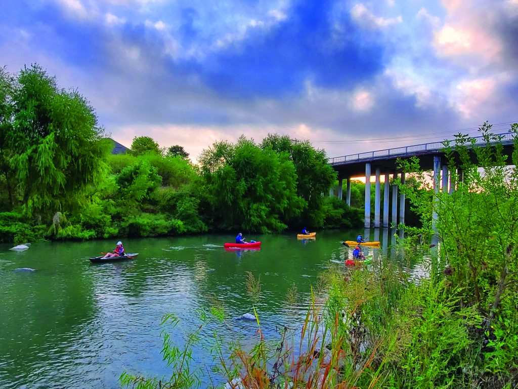 Photo of kayakers paddling in the Mission Reach section of the San Antonio River. They kayakers are in a calm section of river, right near an overpass.