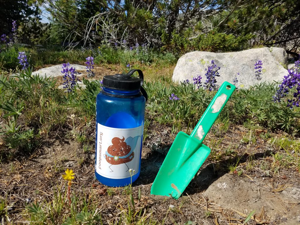A water bottle with a PoopSmart sticker on it sits next to a green hand shovel at Whistler Pass in British Columbia.