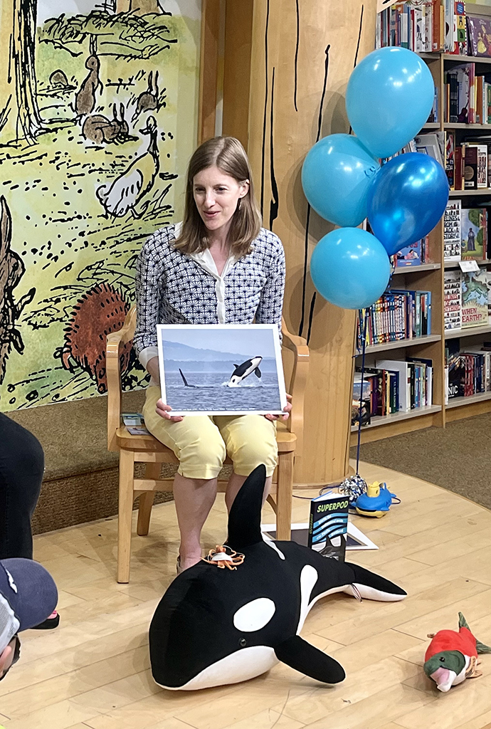Photo of Nora Nickum speaking with kids about her book, "Superpod," at a nearby Barnes & Noble store. Nora is holding a photo of a Southern Resident orca, sitting on a chair, and has a stuffed orca on the ground in front of her.