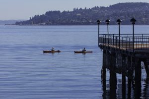 Photo of two kayakers paddling in Commencement Bay, near Tacoma. There's a pier on the right-hand side of the photo, and Brown's Point is in the distance.
