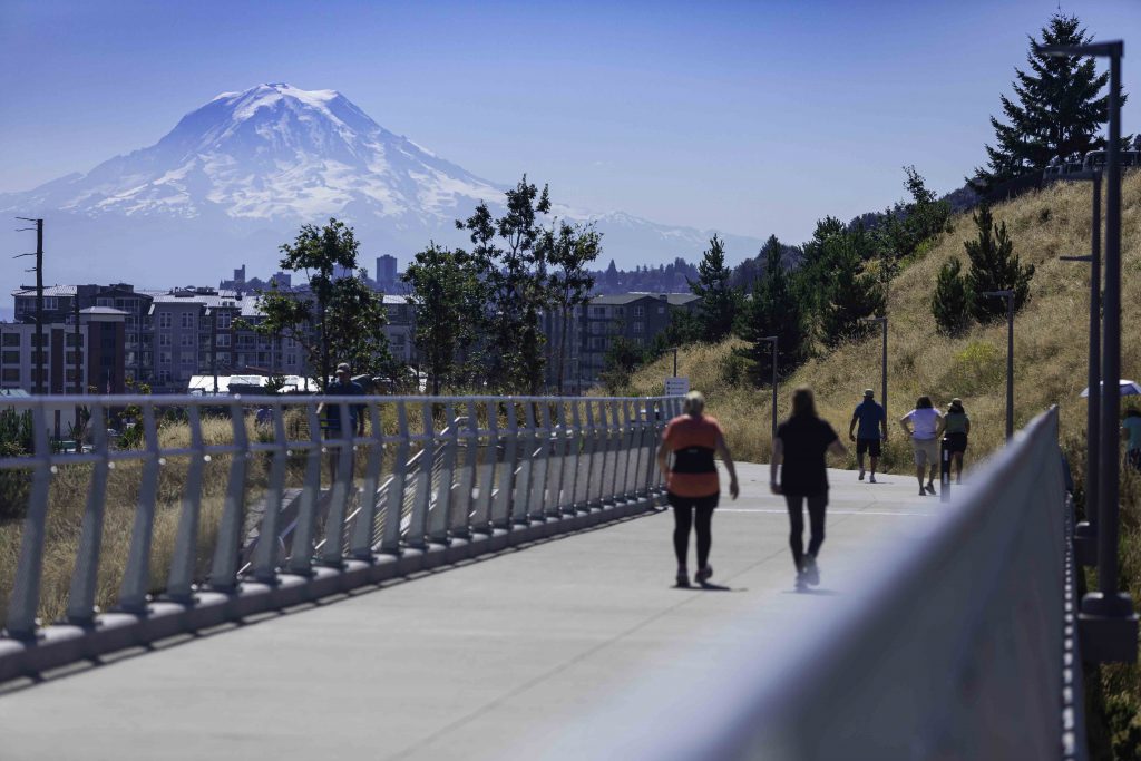 Photo of two people walking along the paved path that leads from Point Defiance Park down to Point Ruston. Mount Rainier is in the background.