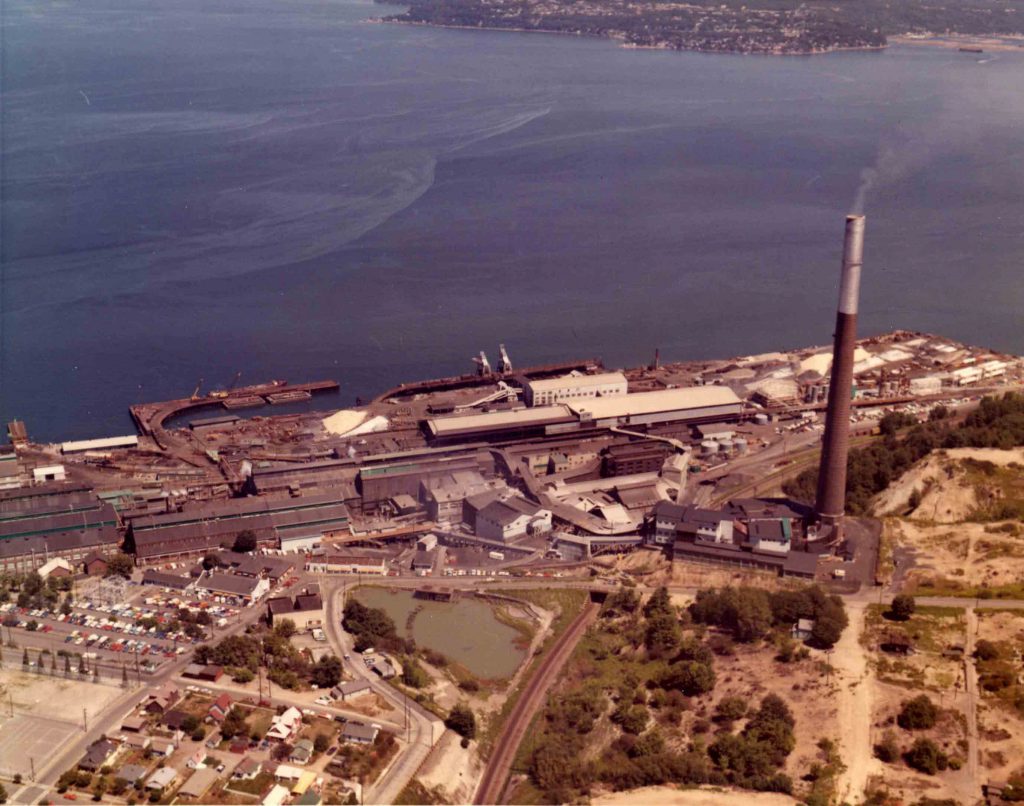 Aerial view of the ASARCO Tacoma smelter site, showing the smokestack towering above the smelter site and Commencement Bay in the background.