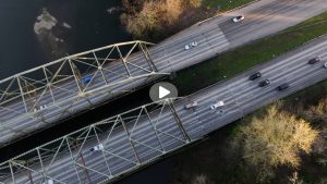 Aerial view of the I-5 bridge over the Nisqually River, with traffic passing over the bridge. A play button is superimposed on the photo to indicate that this story is a playable video.